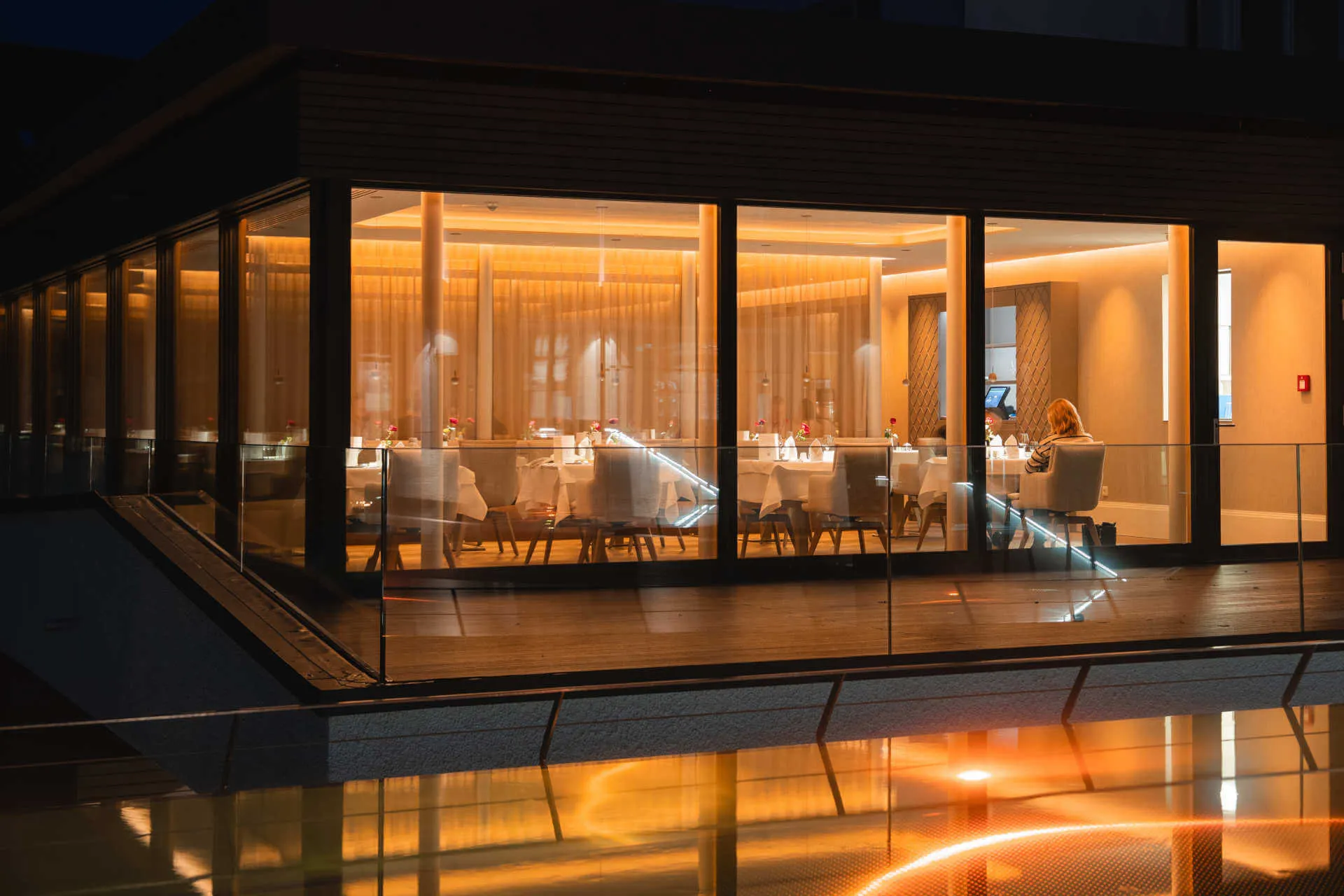 Restaurant mit großen Fenstern, abends warm beleuchtet, Spiegelung im Wasser davor.