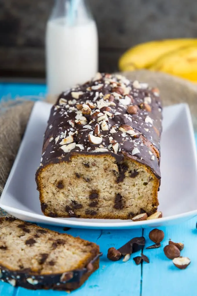 Scheiben von veganem Bananenbrot mit Schokolade und Haselnüssen auf weißem Teller, mit Milchflasche und Bananen im Hintergrund
