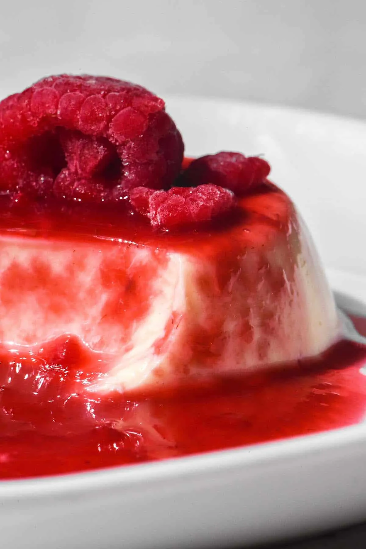 Seitenansicht einer Panna Cotta mit Himbeercoulis und einer gefrorenen Himbeere, angerichtet auf einem weißen Teller vor hellem Hintergrund.