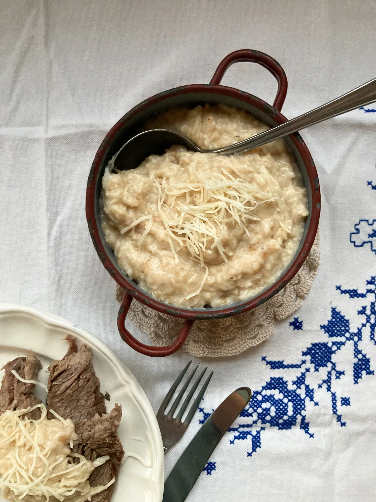 Semmelkren angerichtet in rustikalem Emaille-Topf, neben Teller mit gekochtem Rindfleisch. Die dampfende Semmelkren wird mit einem Löffel serviert und erfreut das Auge mit ihrer cremigen Konsistenz und dem rustikalen Charme.
