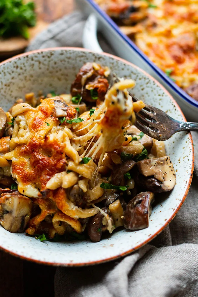 Spätzle-Pilz-Auflauf mit Käsefäden in einer Schüssel. Im Hintergrund ist die Auflaufform zu sehen.