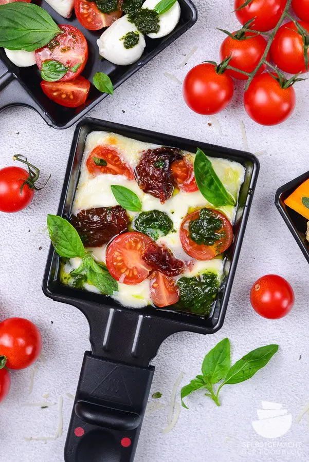Überbackene Caprese im Raclette Pfännchen mit frischen Tomatenscheiben, Mozzarella, Basilikum und Basilikumpesto, eine schnelle vegetarische Raclette Idee.