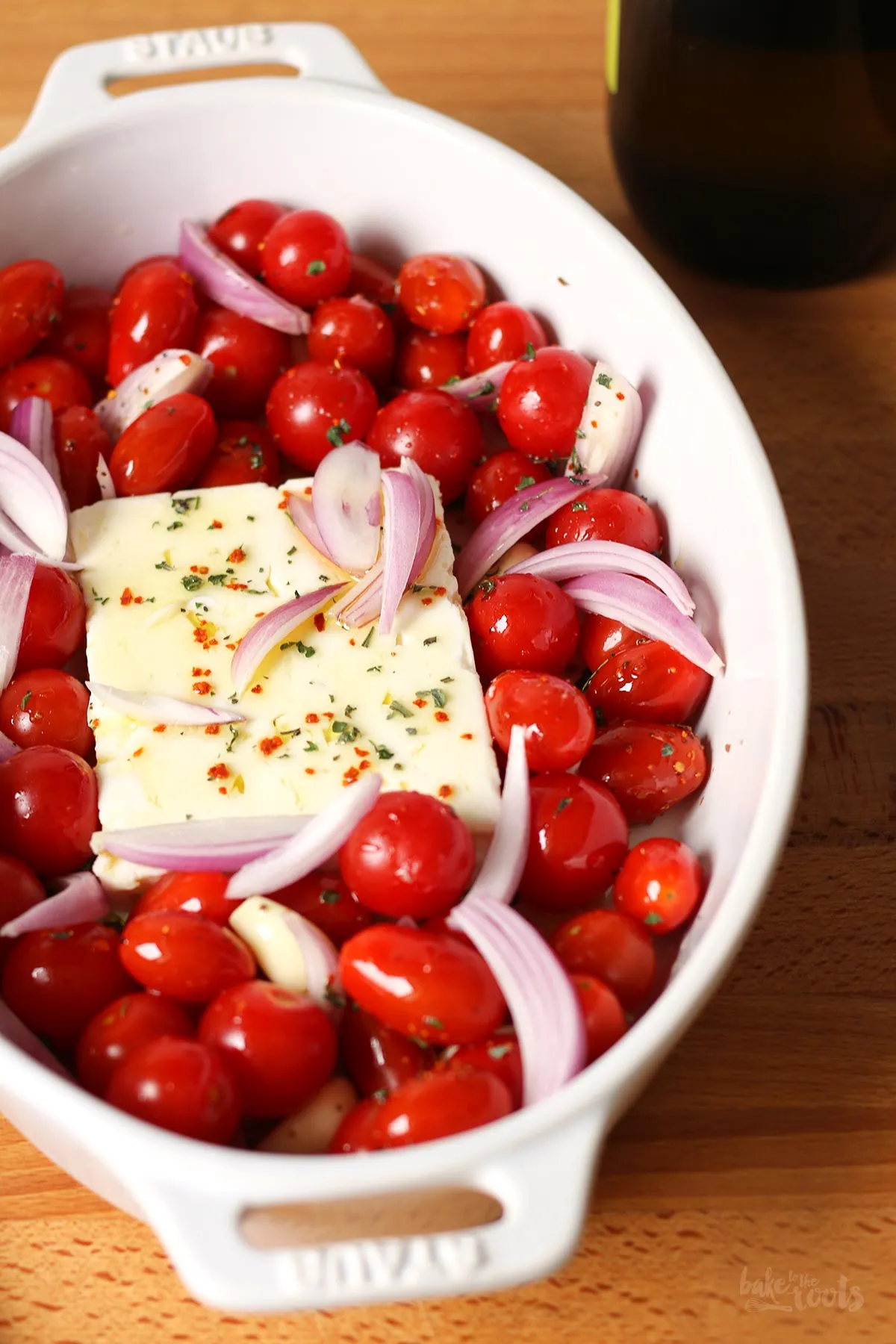 Ungebackener Feta-Block in einer Auflaufform, umgeben von frischen Cherrytomaten, Zwiebeln und Knoblauchzehen, bereit für den Ofen.