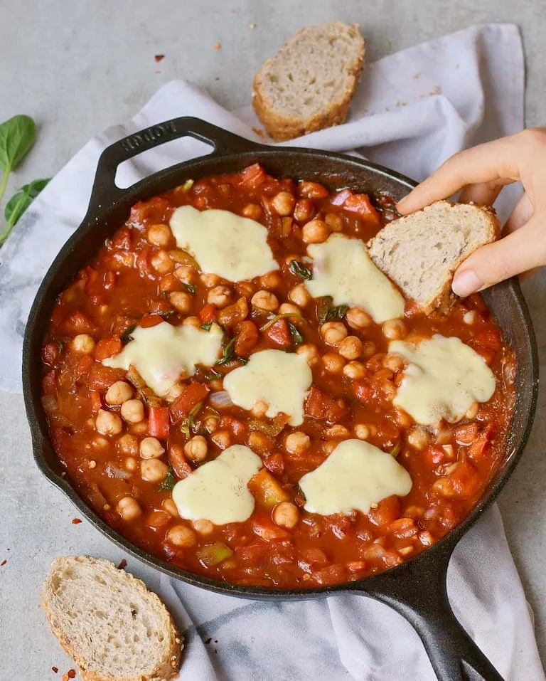 Veganes Kichererbsen-Shakshuka mit viel Gemüse in rauchiger Tomatensoße in einer schwarzen Pfanne, ein nahrhaftes Mittagessen.