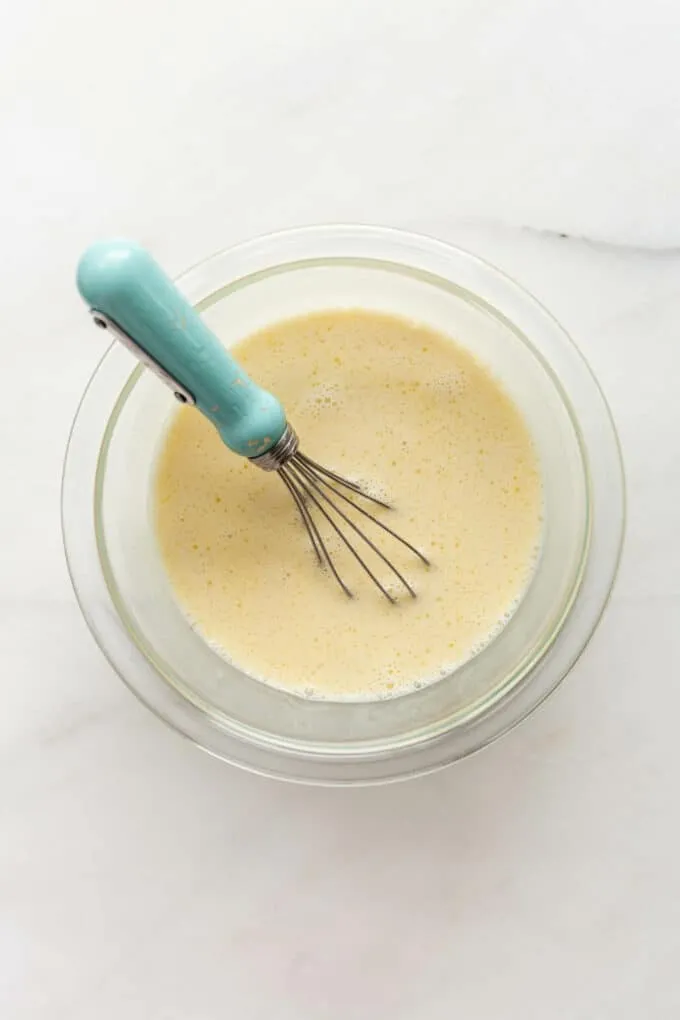 Verquirlte Eier und Milch in einer transparenten Schüssel mit einem blauen Schneebesen.