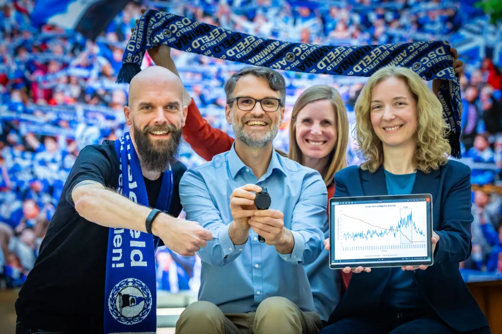 Vier Personen (Jens Franzke, Christian Deutscher, Julia Bömer und Christiane Fuchs) halten Smartwatches und ein Tablet vor einem unscharfen Hintergrundbild mit Fans im Stadion.