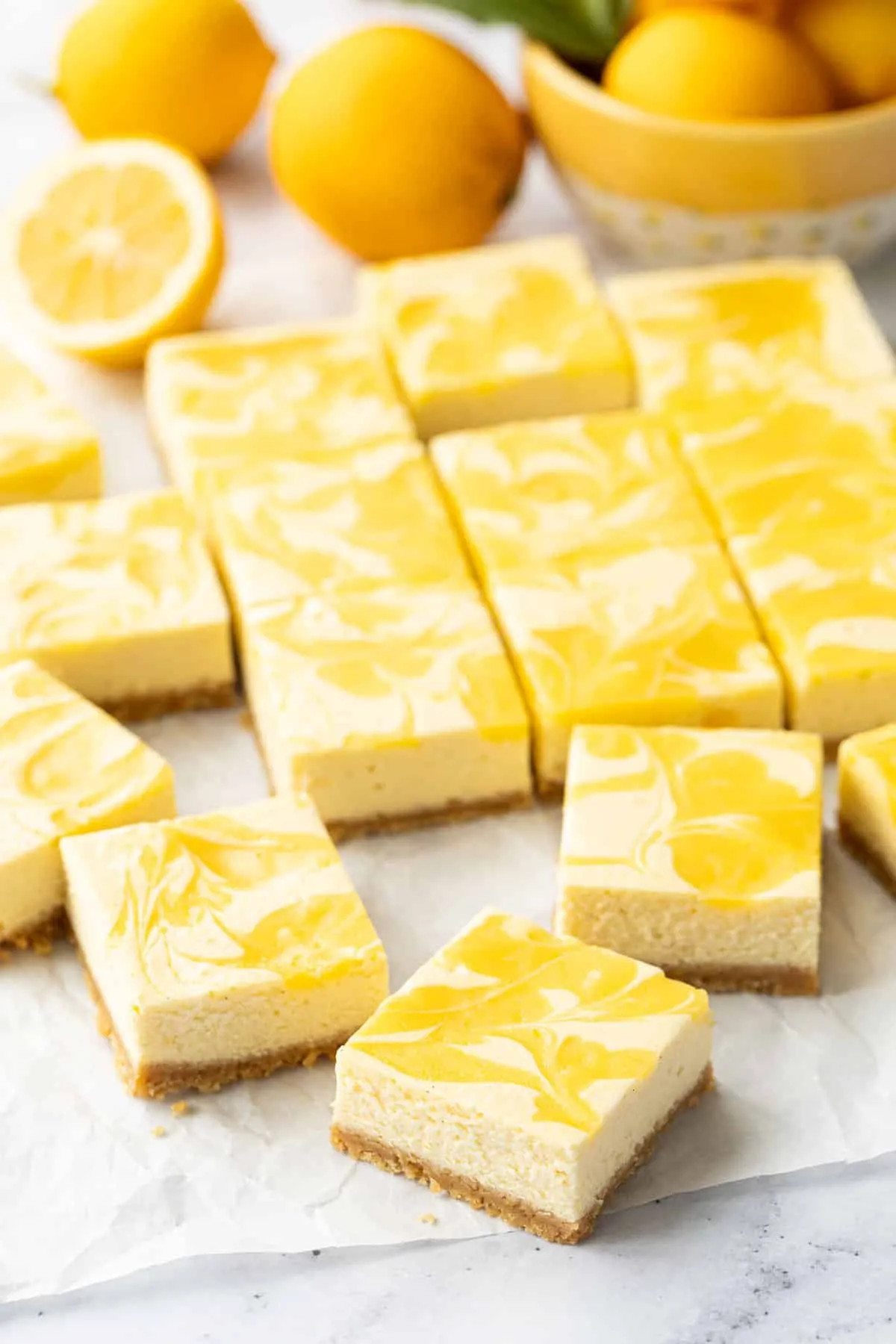 Zitronen-Käsekuchenriegel mit Lemon Curd, unregelmäßig auf zerknittertem Backpapier arrangiert, frische Zitronen im Hintergrund.