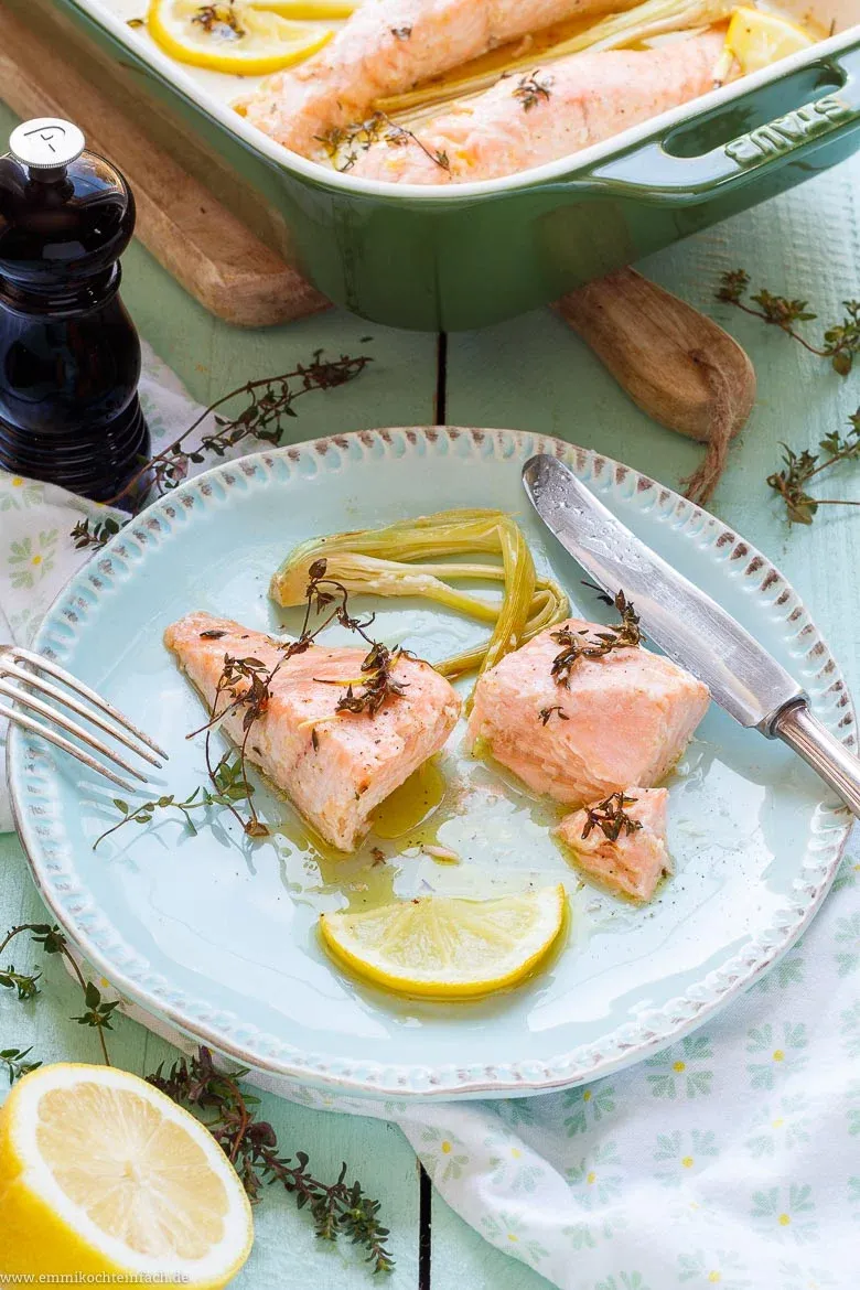 Zitronen-Lachsfilets mit frischen Zitronenscheiben, angedrücktem Knoblauch und Thymianzweigen in einer Auflaufform, übergossen mit Olivenöl-Dressing.