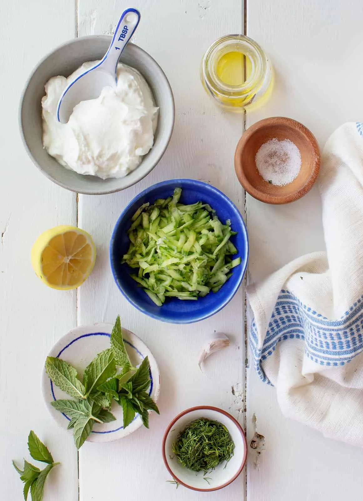 Zutaten für selbstgemachtes Tzatziki, darunter Gurke, griechischer Joghurt, Dill, Minze, Knoblauch und Zitrone, auf einem Schneidebrett arrangiert.