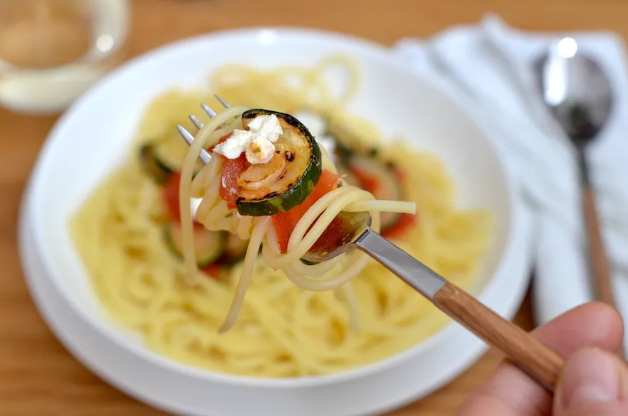 Zutaten für Spaghetti mit Zucchini, Tomaten und Feta auf einem Holztisch arrangiert. Zu sehen sind frische Zucchini, Tomaten, Knoblauchzehen, Olivenöl und eine Packung Feta-Käse.