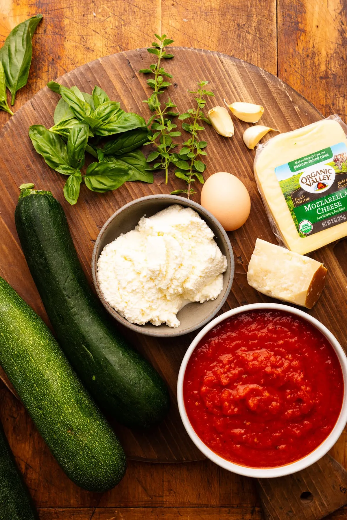 Zutaten für Zucchini Lasagne auf einem Holzbrett angerichtet, darunter Zucchini, Parmesankäse, Mozzarella, Ricotta, Knoblauch und frische Kräuter.