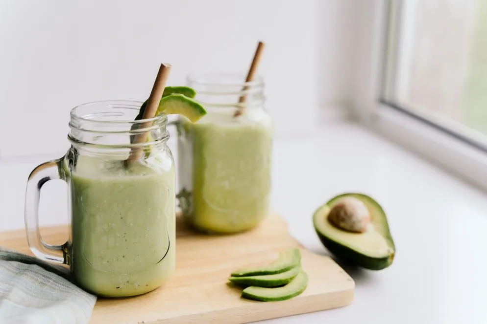 Zwei Gläser mit grünem Avocado-Smoothie und Strohhalmen auf einem Holzbrett, daneben eine aufgeschnittene Avocado.