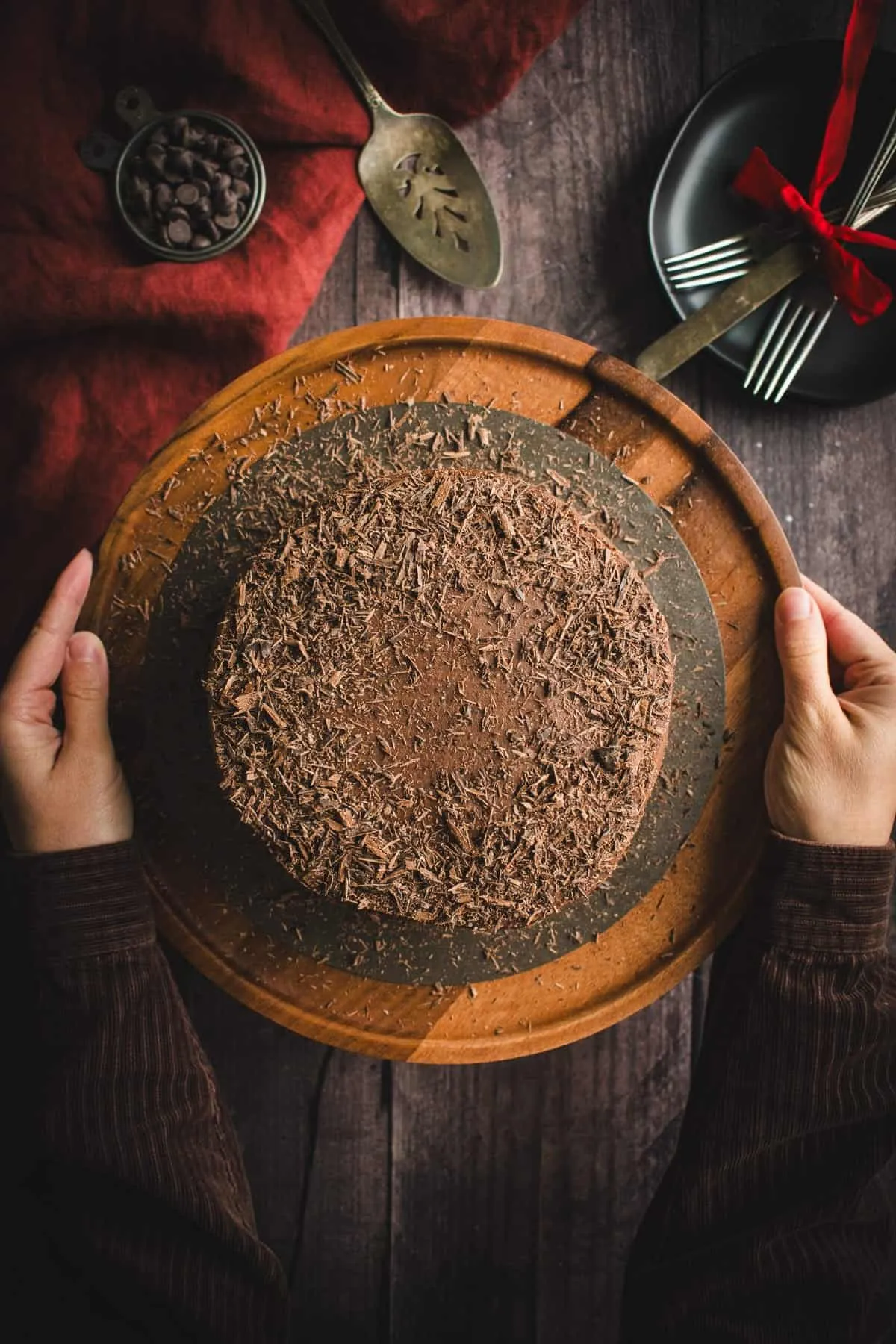 Zwei Hände halten einen hölzernen Tortenständer mit einer schokoladenfrosted Torte darauf.