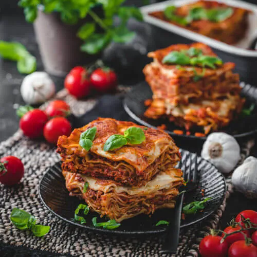 Zwei kunstvoll geschichtete Stücke Lasagne, präsentiert auf eleganten schwarzen Tellern auf einem dunklen Untersetzer. Dekoriert mit Basilikum, Knoblauch und Tomaten.