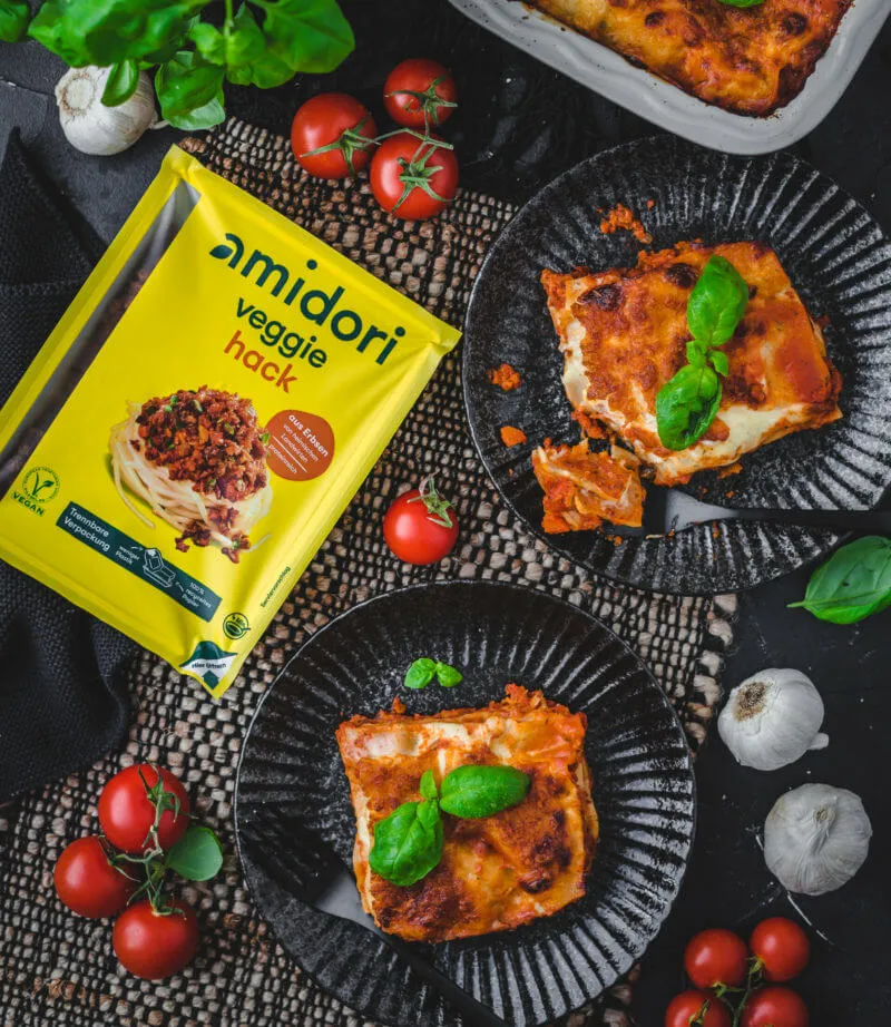 Zwei schwarze Teller mit jeweils einer Portion vegetarischer Lasagne darauf, garniert mit Basilikumblättern. Daneben eine Packung veganer Hackfleischersatz in der Schale.