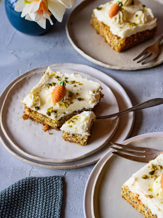 3 Stück Karotten-Mohn-Blechkuchen mit Mascarpone-Topping auf Tellern, beim mittleren Stück teilt eine Gabel eine Ecke ab, von schräg vorne aufgenommen