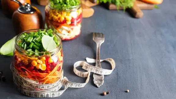 Abnehmen mit der richtigen Ernährung: Glas gefüllt mit Gemüse. Um das Glas und eine Gabel ist ein Maßband gewickelt.