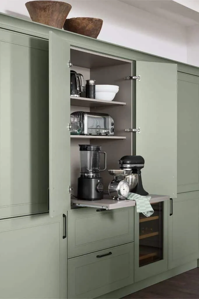 Ausgezogener Tablarauszug in einem grünen Küchenschrank mit Küchengeräten wie Mixer, Küchenmaschine und Waage, darüber offene Regalböden mit weiteren Geräten und Geschirr.