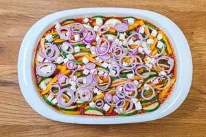 Der Kritharaki-Auflauf wird vor dem Backen mit den restlichen Fetawürfeln und roten Zwiebelringen belegt, für zusätzliche Würze und Röstaromen