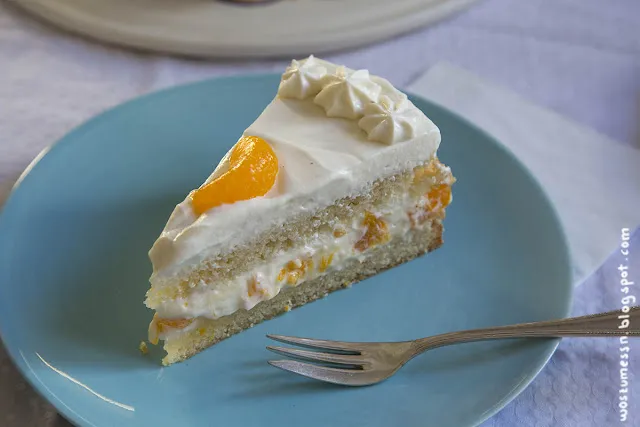 Ein großes Stück vegane Käse-Sahne-Torte mit Mandarinen, angeschnitten auf einem Teller, zeigt die cremigen Schichten und den Biskuitboden.