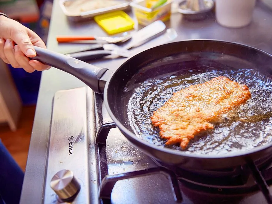 Ein Wiener Schnitzel wird in einer Pfanne mit viel heißem Fett gebraten, wobei das Fett über das Schnitzel gegossen wird.