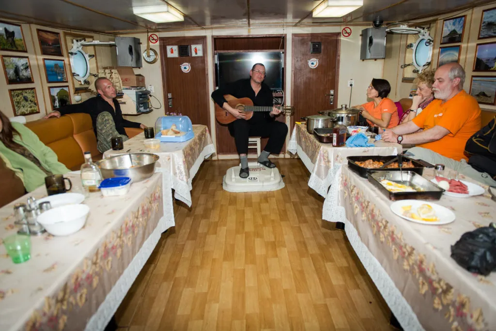 Eine Gruppe von Menschen sitzt um ein Lagerfeuer und entspannt sich in einer ruhigen Abendstimmung am Wasser, mit einer dunklen Bergkulisse im Hintergrund.