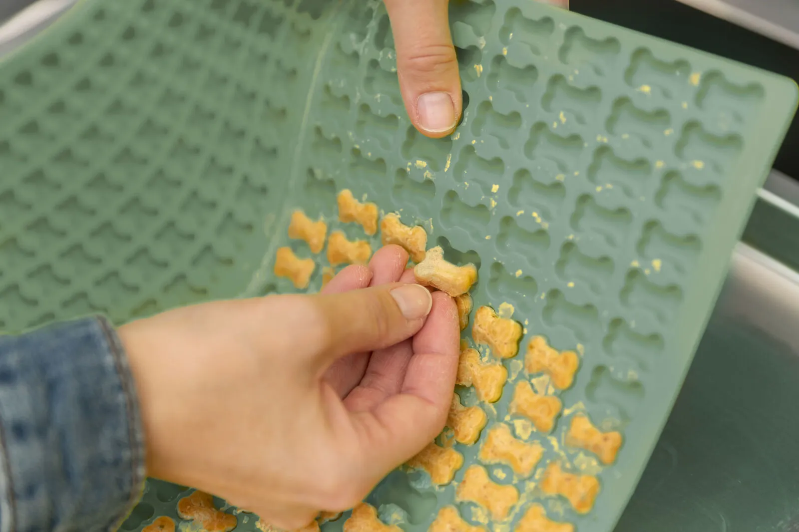 Eine Hand löst spielend leicht goldbraun gebackene Hundekekse in Knochenform aus einer blauen Silikon-Backmatte.