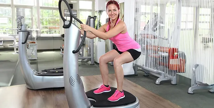 Frau im Fitnessstudio macht Kniebeuge auf Vibrationsplatte zur Kräftigung von Beinen und Gesäß.