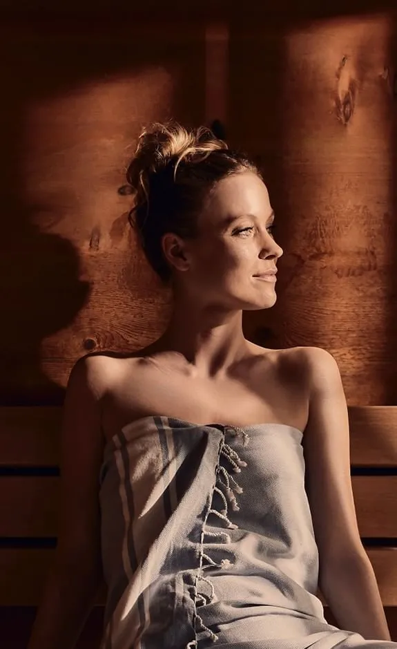 Frau in einem Bademantel auf einer hölzernen Saunabank sitzend, mit warmem Licht beleuchtet.