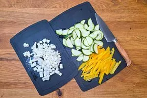 Geschnittene gelbe Paprika, Zucchinischeiben und gewürfelten Feta auf einem Schneidebrett für den griechischen Nudelauflauf