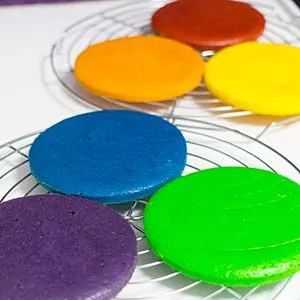 Mehrere ausgekühlte, runde Kuchenböden in verschiedenen Regenbogenfarben liegen bereit
