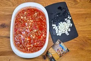 Ungekochte Kritharaki-Nudeln und die Hälfte der Fetawürfel werden mit der würzigen Hackfleischsoße in einer großen Auflaufform vermengt