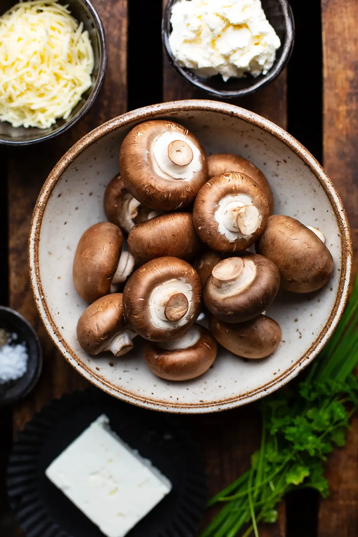 Zutaten für gefüllte Champignons: Braune Pilze, Feta, Frischkäse, Kräuter und geriebener Käse auf dunklem Holz