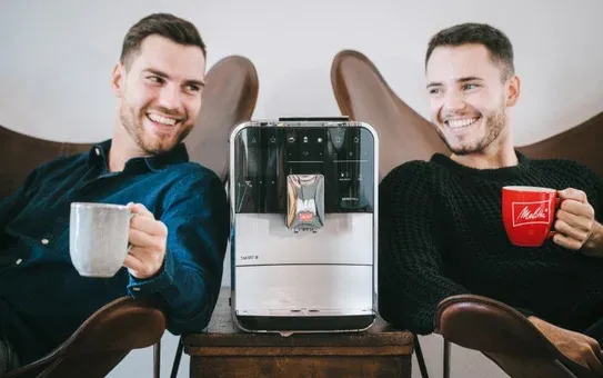 Zwei Männer genießen Kaffee mit einer silbernen Melitta CI Touch zwischen sich