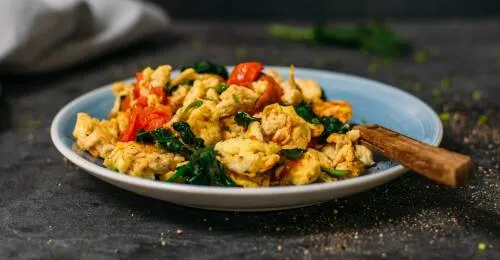 Blauer Teller gefüllt mit Rührei, Tomaten und Spinat, vor dunkelgrauem Hintergrund. Von der Seite fotografiert.
