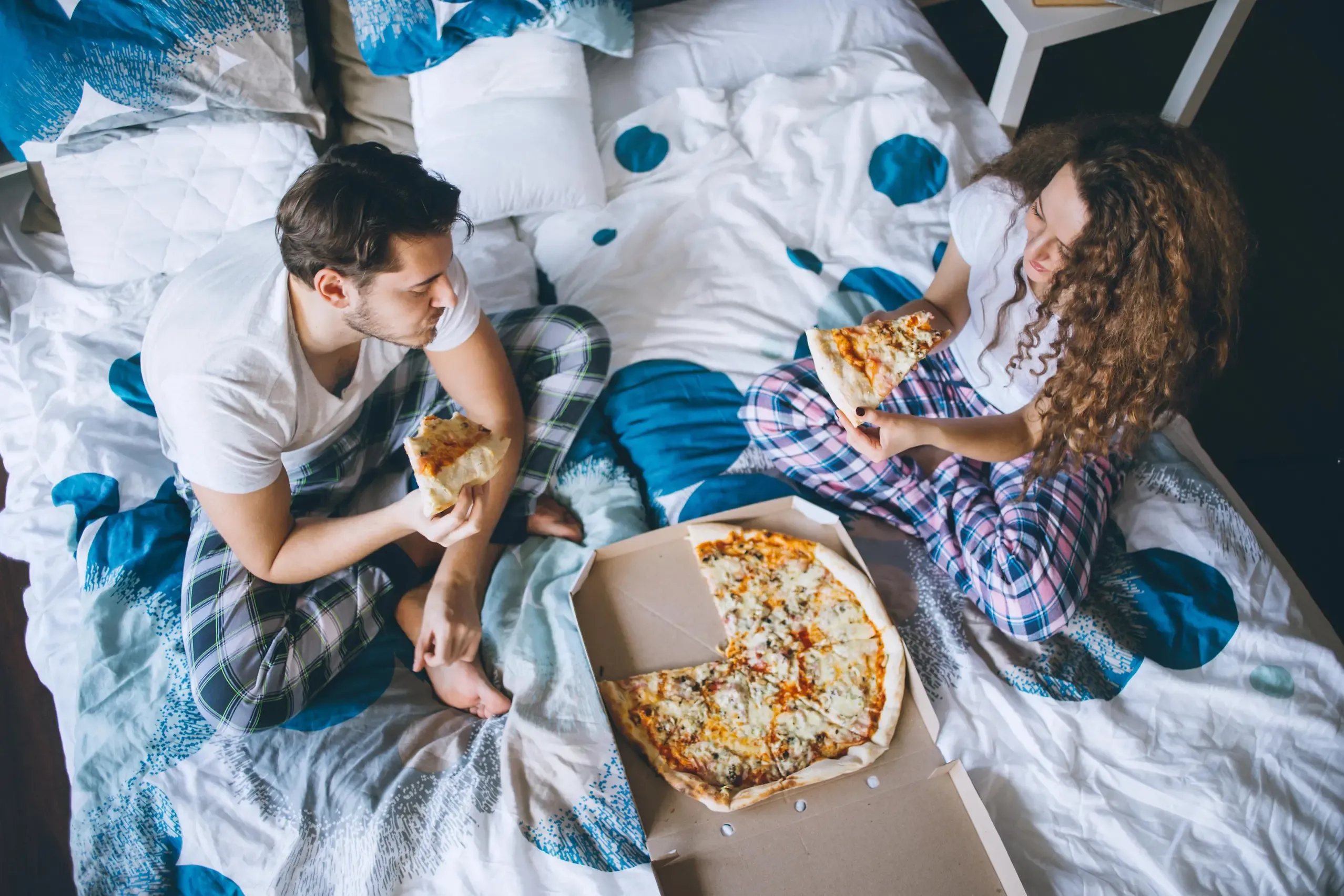 Zwei Personen sitzen auf dem Bett und essen genüsslich eine Pizza