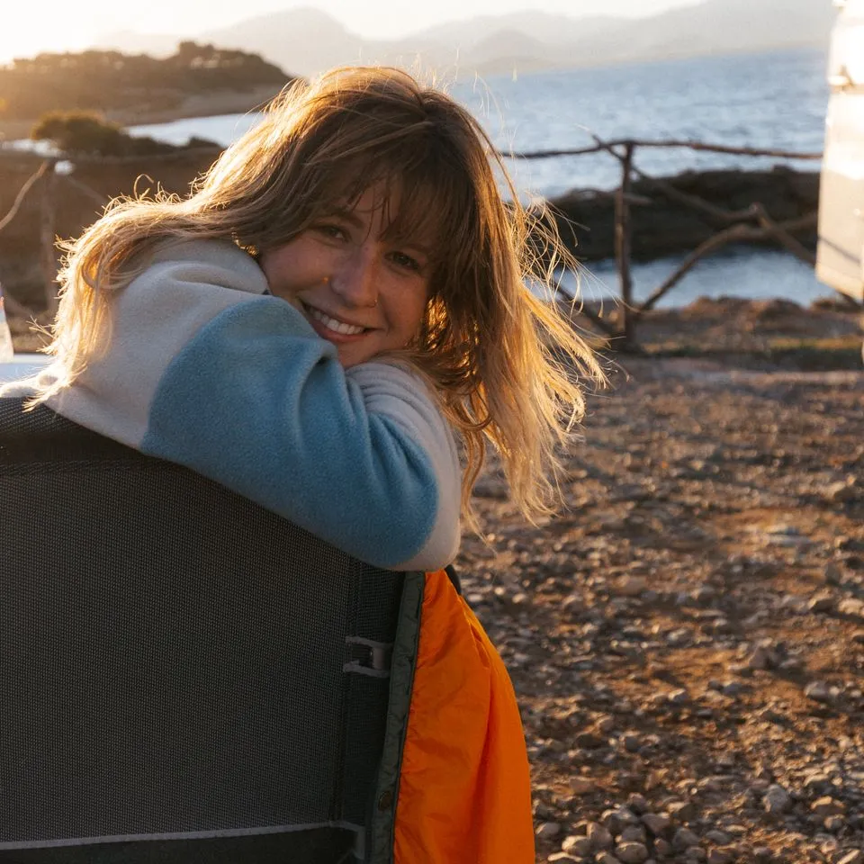 Warme Reiseziele April: Junge Frau sitzt lachend neben einem Camper am Meer in der Sonne