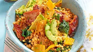 Warmer Linsensalat mit roten Linsen, Avocadostreifen, gelbem Gemüse und gebratenen Tomaten in einer türkisfarbenen Keramikschüssel.