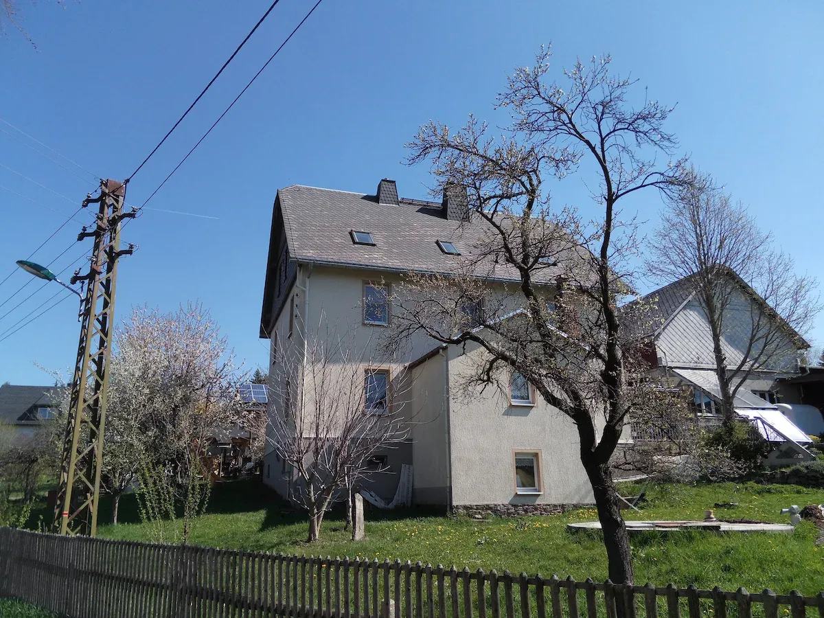 Außenansicht eines dreistöckigen Gebäudes, umgeben von Grün und Bäumen. Ein Holzzaun begrenzt das Grundstück, während ein klarer blauer Himmel die ländliche Umgebung betont. Das Gebäude verfügt über mehrere Fenster und ein Giebeldach.