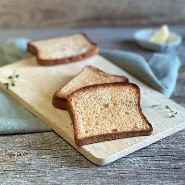 Drei Scheiben Toastbrot auf einem Holzbrett. Im Hintergrund ein grünes Tuch und eine Schale mit Butter.
