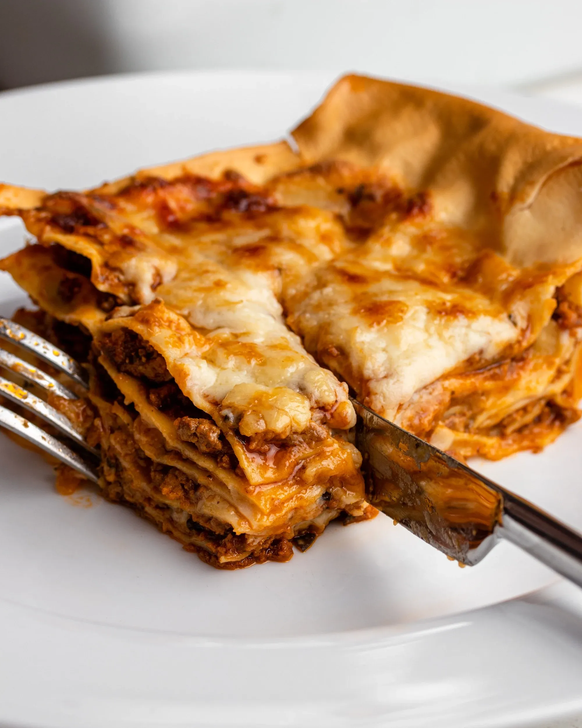 Ein Stück Lasagne auf einem Teller mit Messer und Gabel, das durch die Schichten schneidet