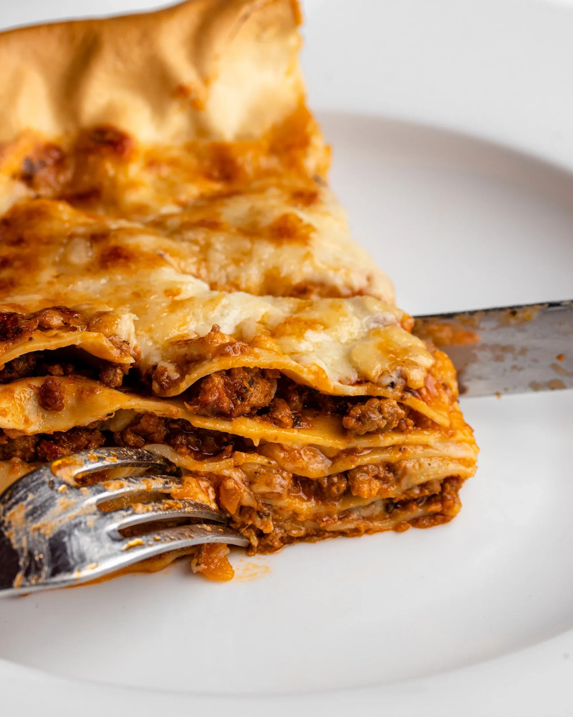 Ein Stück Lasagne auf einem weißen Teller, das mit Messer und Gabel aufgeschnitten wird