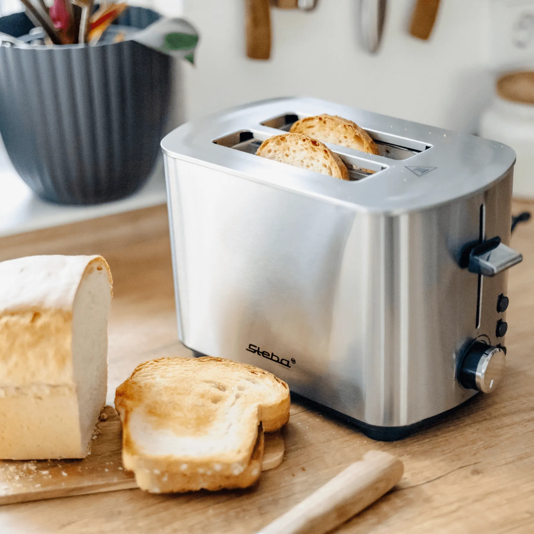 Ein Toaster von Steba aus Edelstahl mit zwei Schlitzen, neben frischem Brot und zwei gerösteten Scheiben auf einem Holzbrett.
