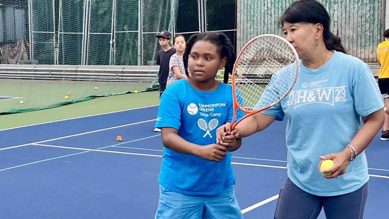 Ein Trainer leitet ein Kind mit Tennisschläger auf einem Außenplatz an, umgeben von Bällen