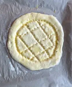 Eine Hand drückt Muster in ein vorbereitetes Fladenbrot auf einem Backblech, während es mit Eigelb-Butter-Mischung bestrichen wird
