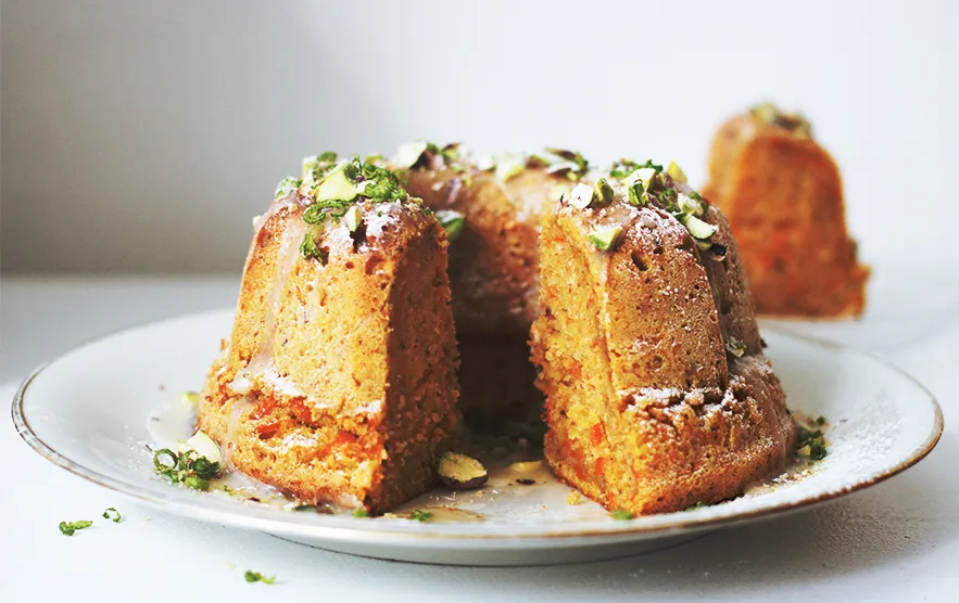 Eine Hand hält ein Stück Karottenkuchen mit Guss und Pistazien
