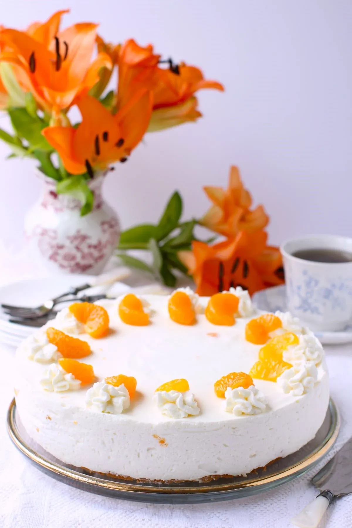 Eine Mandarinen-Sahnetorte auf dem Tisch mit einer Vase orangefarbener Lilien dahinter.