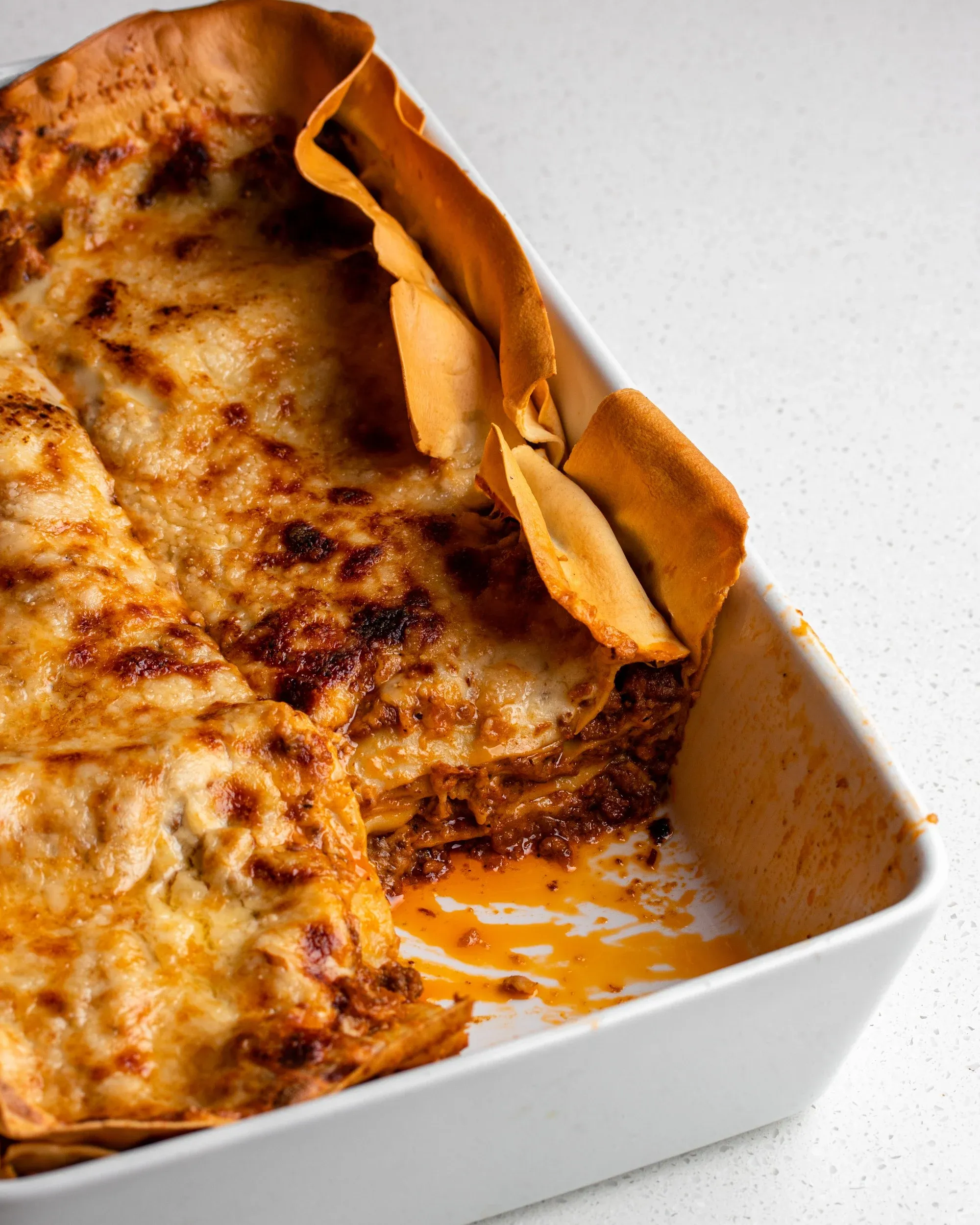 Eine weiße Keramikauflaufform mit einer gekochten Lasagne. Goldbraune Oberseite und knusprige Seiten. Ein Stück wurde herausgeschnitten.