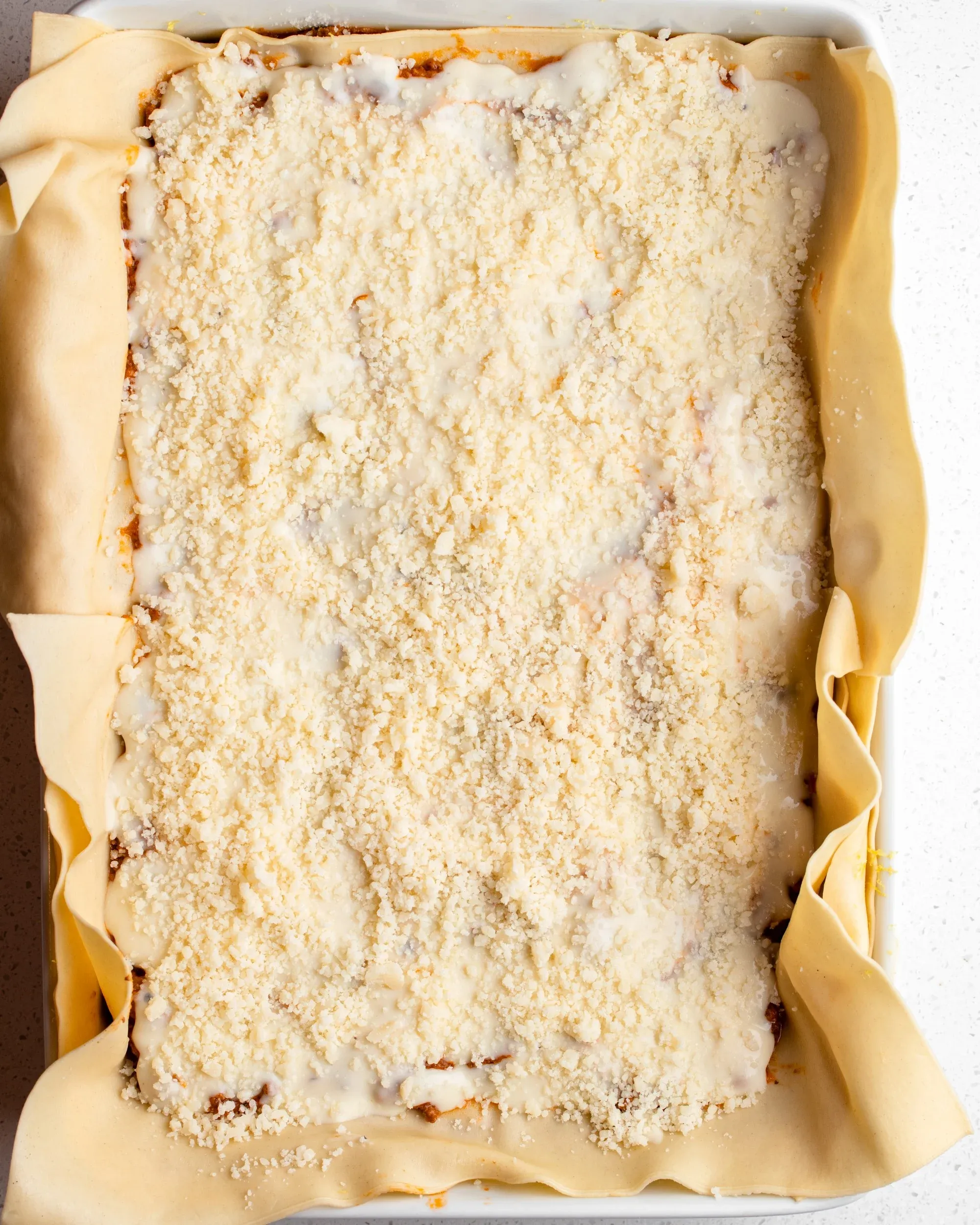 Fertig zusammengebaute Lasagne bereit zum Backen. Die Oberseite ist mit Parmesankäse bedeckt und die Nudelplatten hängen über die Seiten der Form.