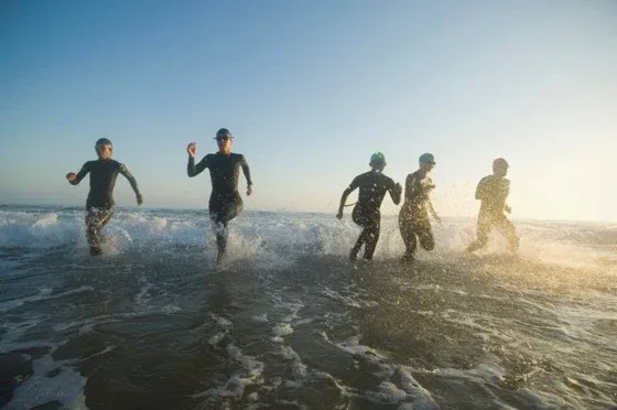 Fünf Personen in Schwimmanzügen laufen durch das Meerwasser bei Sonnenaufgang, was auf einen Triathlon hindeutet.